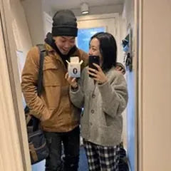 A couple taking a mirror selfie in their hallway; she holds up one open box.