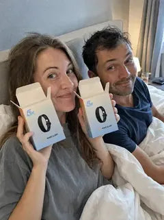 A couple in bed: she holds two open boxes up beside her face while he smiles next to her.