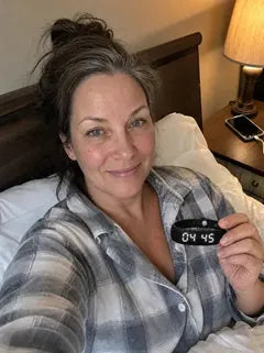A wife in plaid pyjamas, propped up in bed, holding up the band, its screen lit at 04:45.