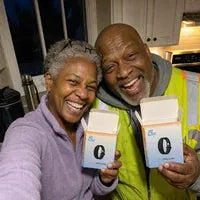 A couple laughing in their kitchen, each holding an open 5AM Solo Wake Alarm box; he is in a hi-vis vest.
