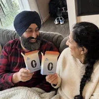 A couple on the sofa under a blanket, smiling at each other, each holding an open box.