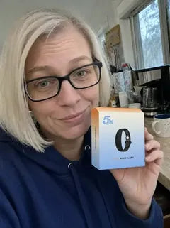 A wife in glasses and a blue hoodie, in her kitchen, holding the 5AM Solo Wake Alarm box.