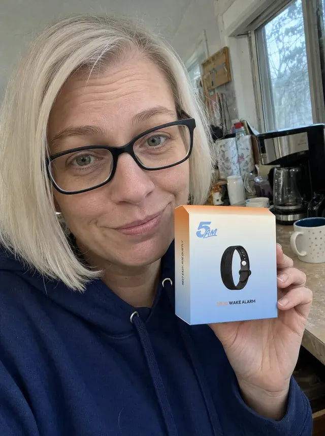 A wife in glasses and a blue hoodie, in her kitchen, holding the 5AM Solo Wake Alarm box.