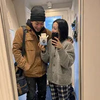 A couple taking a mirror selfie in their hallway; she holds up one open box.