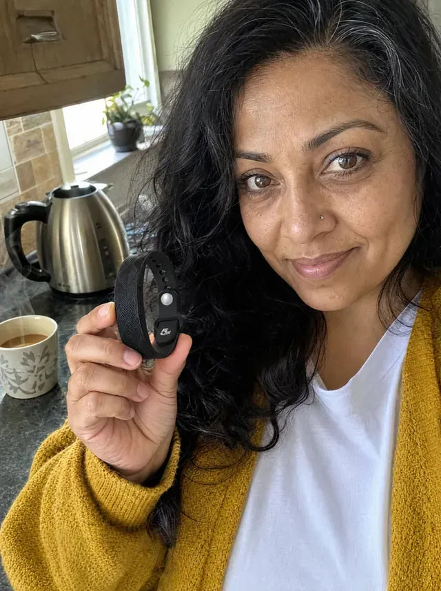 A wife in a yellow cardigan in her kitchen, a cup of chai beside her, holding up the band.
